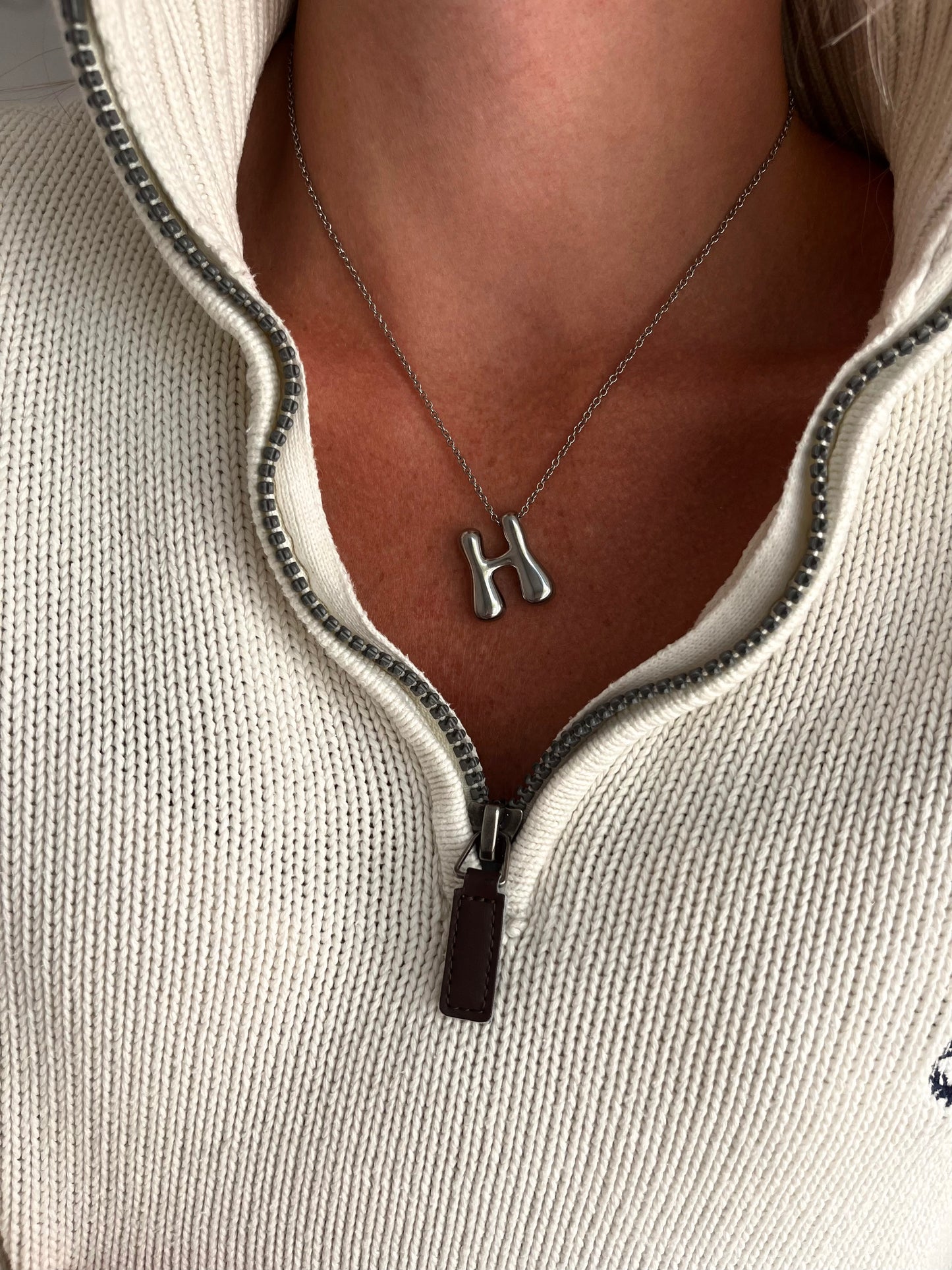 Necklace with a 'H' pendant worn by a person in a light gray zip-up sweater.