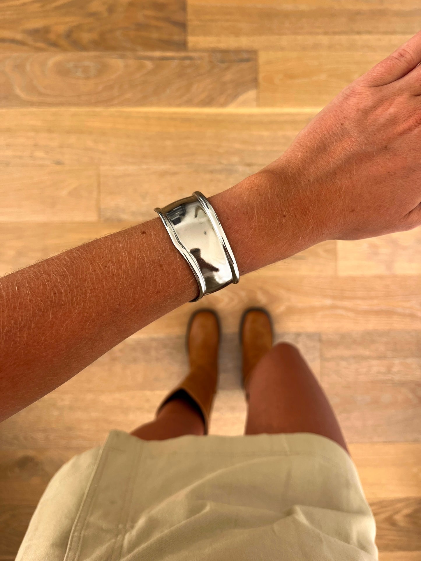 A wrist wearing the Arc sculptural open bangle in polished stainless steel, photographed from above with a beige skirt and brown boots in the background.