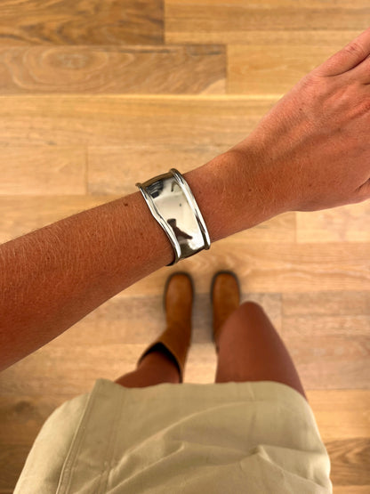 A wrist wearing the Arc sculptural open bangle in polished stainless steel, photographed from above with a beige skirt and brown boots in the background.