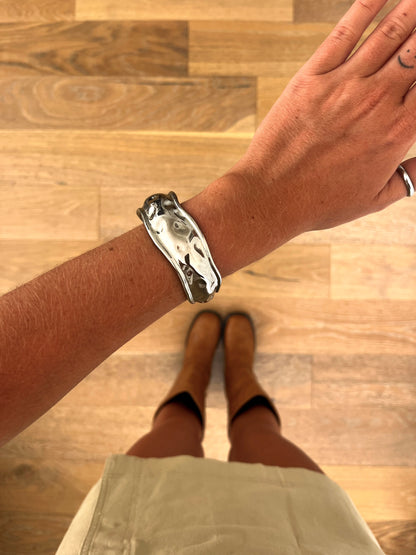 A wrist wearing the Astra sculptural stainless steel bangle, photographed from above with a beige skirt and brown boots in the background.