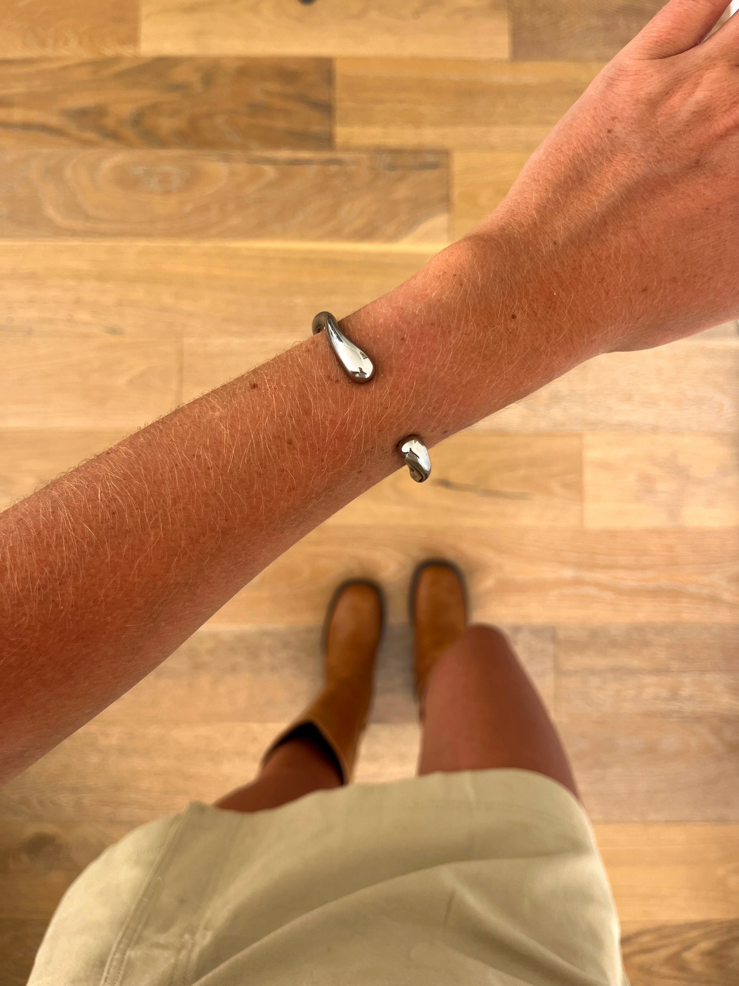 A wrist wearing the Axis sculptural cuff bangle in polished stainless steel, photographed from above with a beige skirt and brown boots in the background.