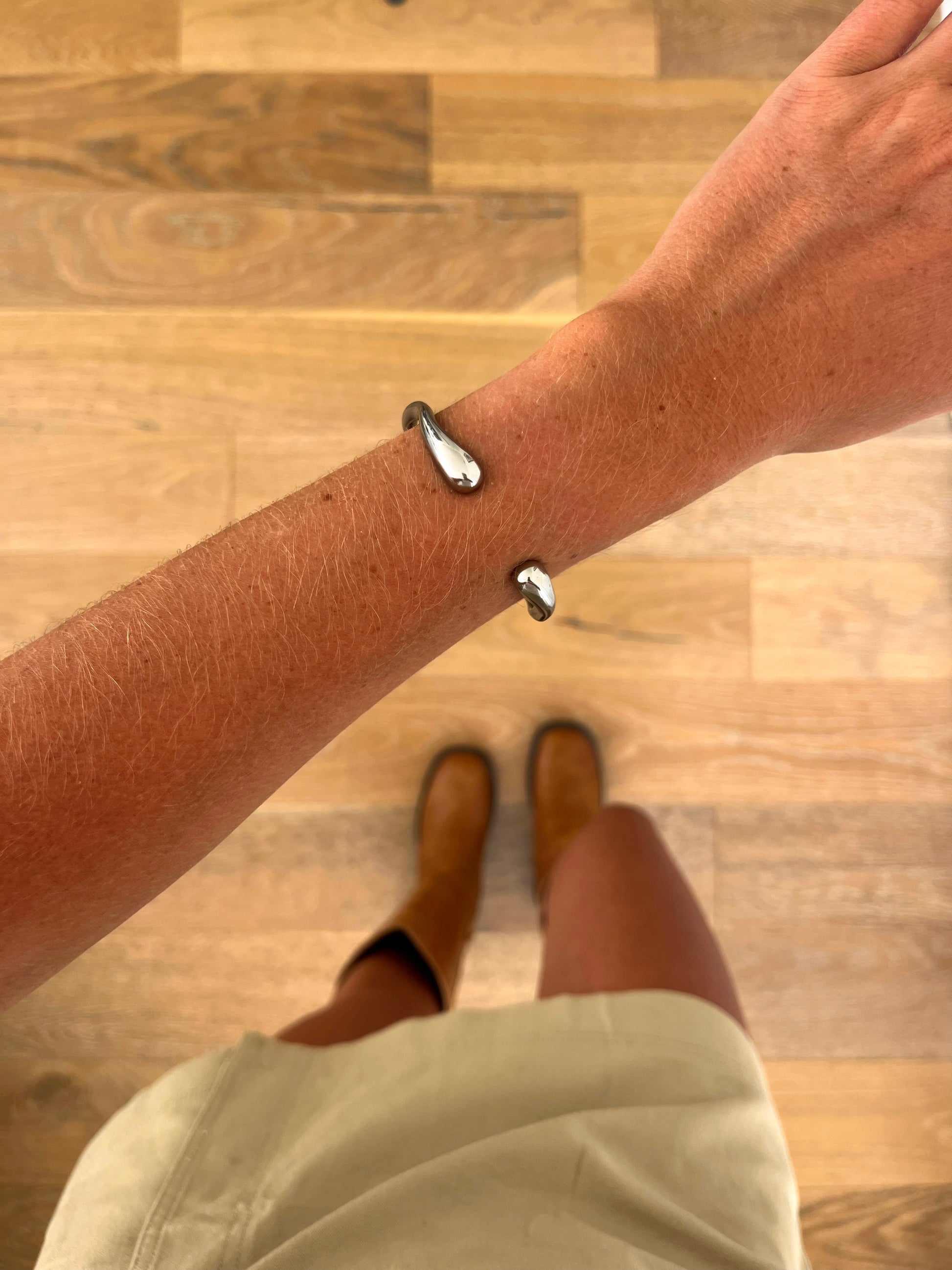 A wrist wearing the Axis sculptural cuff bangle in polished stainless steel, photographed from above with a beige skirt and brown boots in the background.