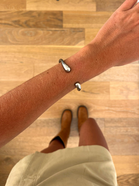 A wrist wearing the Axis sculptural cuff bangle in polished stainless steel, photographed from above with a beige skirt and brown boots in the background.