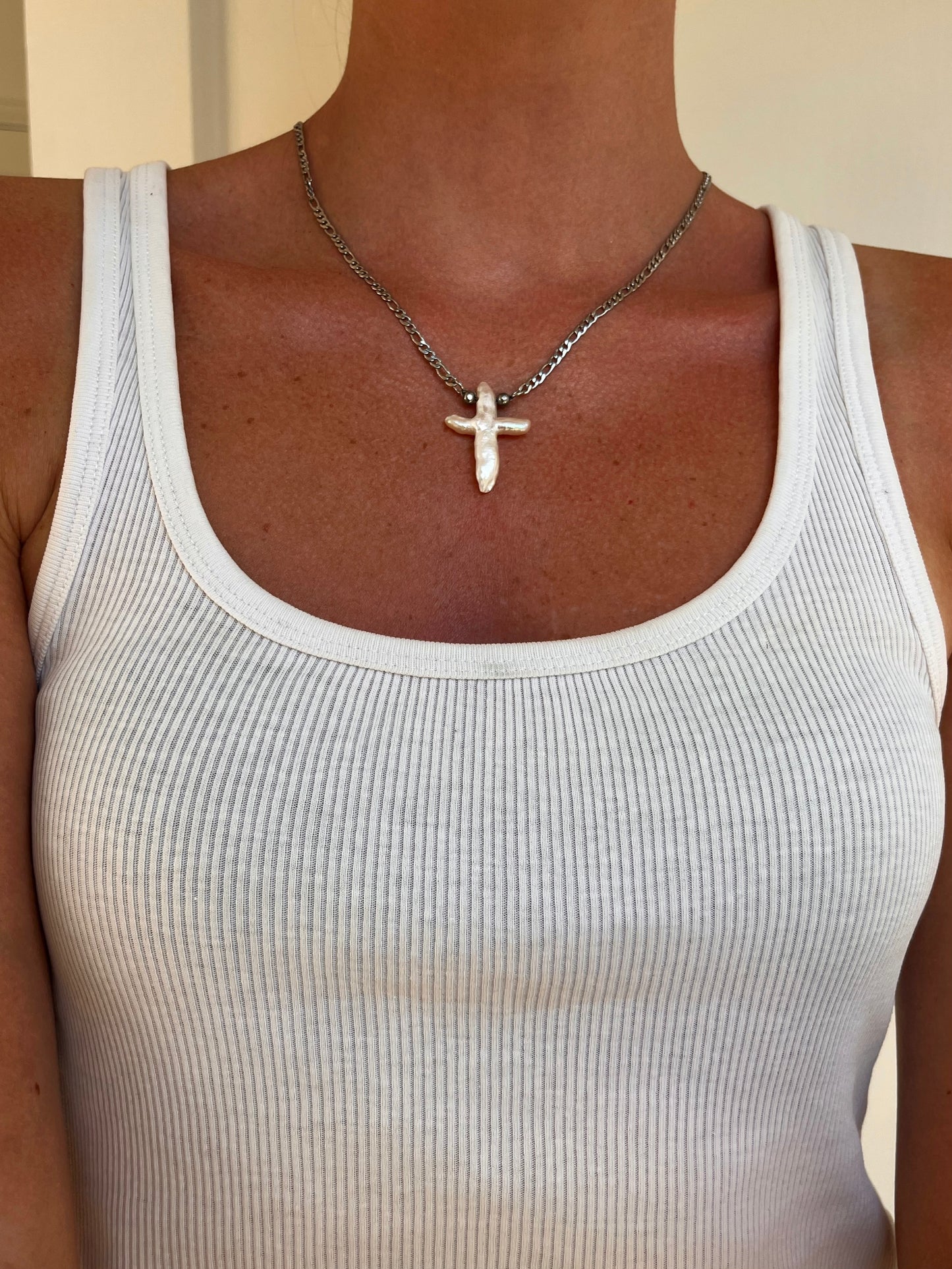 A close-up of a person wearing the crux necklace a silver cross freshwater pendant necklace on a silver chain, paired with a white ribbed tank top.