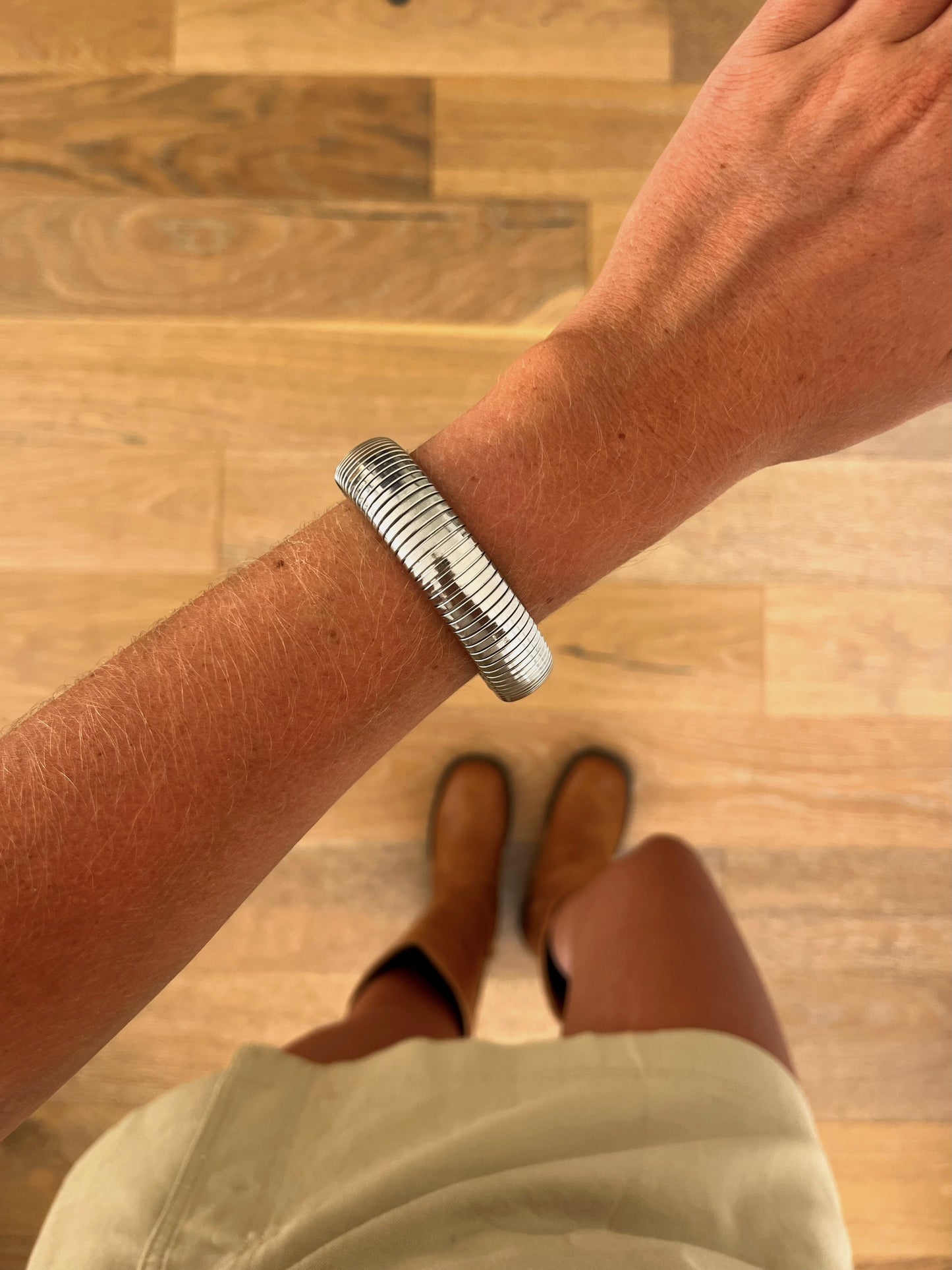 A wrist wearing the Current sculptural stainless steel bangle, photographed from above with a beige skirt and brown boots in the background.