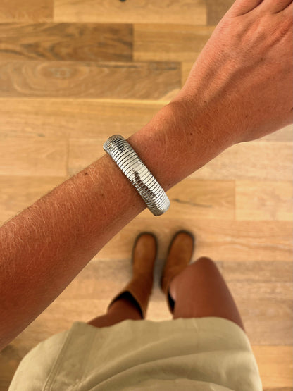 A wrist wearing the Current sculptural stainless steel bangle, photographed from above with a beige skirt and brown boots in the background.
