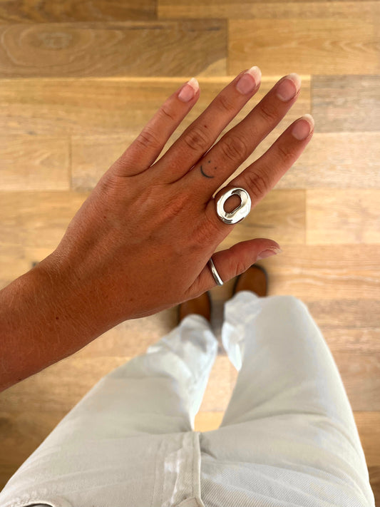 A hand wearing the Dela Compact sculptural open form ring in polished stainless steel, photographed from above with white denim and brown boots in the background.