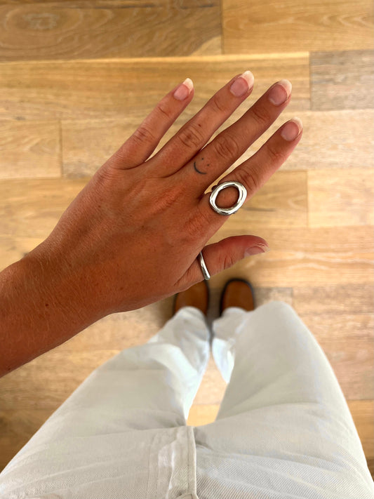 A hand wearing the Doma I sculptural open oval ring in polished stainless steel, photographed from above with white denim and brown boots in the background.