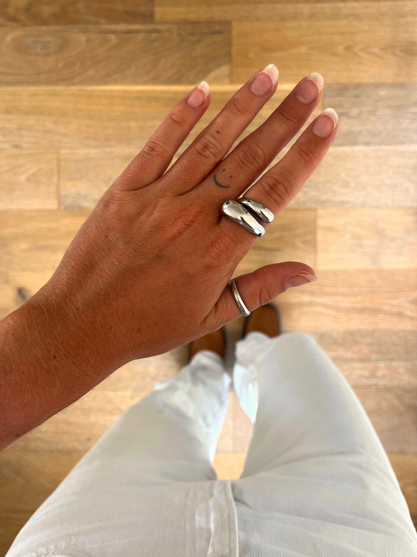 A hand wearing the Dome Arc open sculptural dome ring in polished stainless steel, photographed from above with white denim and brown boots in the background.