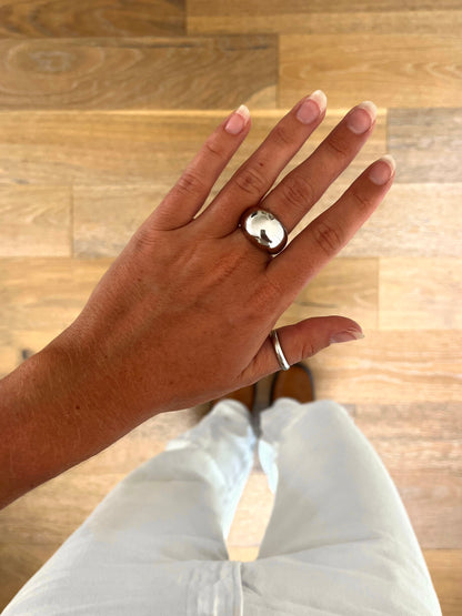 A hand wearing the Dome II rounded dome ring in polished stainless steel, photographed from above with white denim and brown boots in the background.