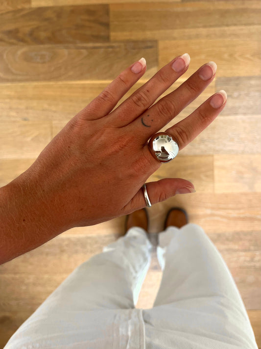 A hand wearing the Dome III bold sculptural dome ring in polished stainless steel, photographed from above with white denim and brown boots in the background.