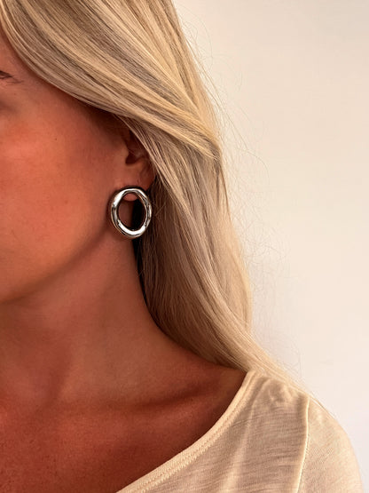 A person wearing the Domena sculptural oval hoop earrings in polished stainless steel.