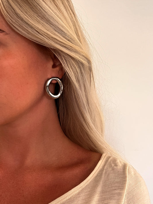 A person wearing the Domena sculptural oval hoop earrings in polished stainless steel.
