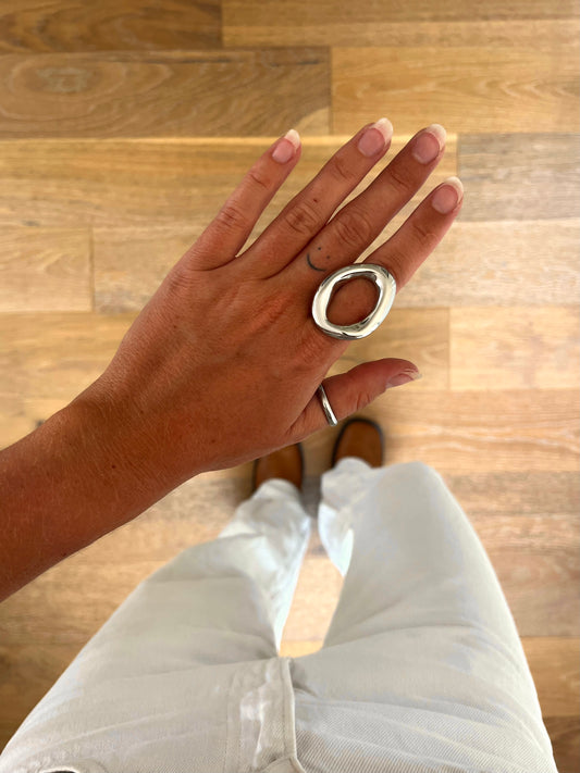 A hand wearing the Domena II sculptural open oval ring in polished stainless steel, photographed from above with white denim and brown boots in the background.