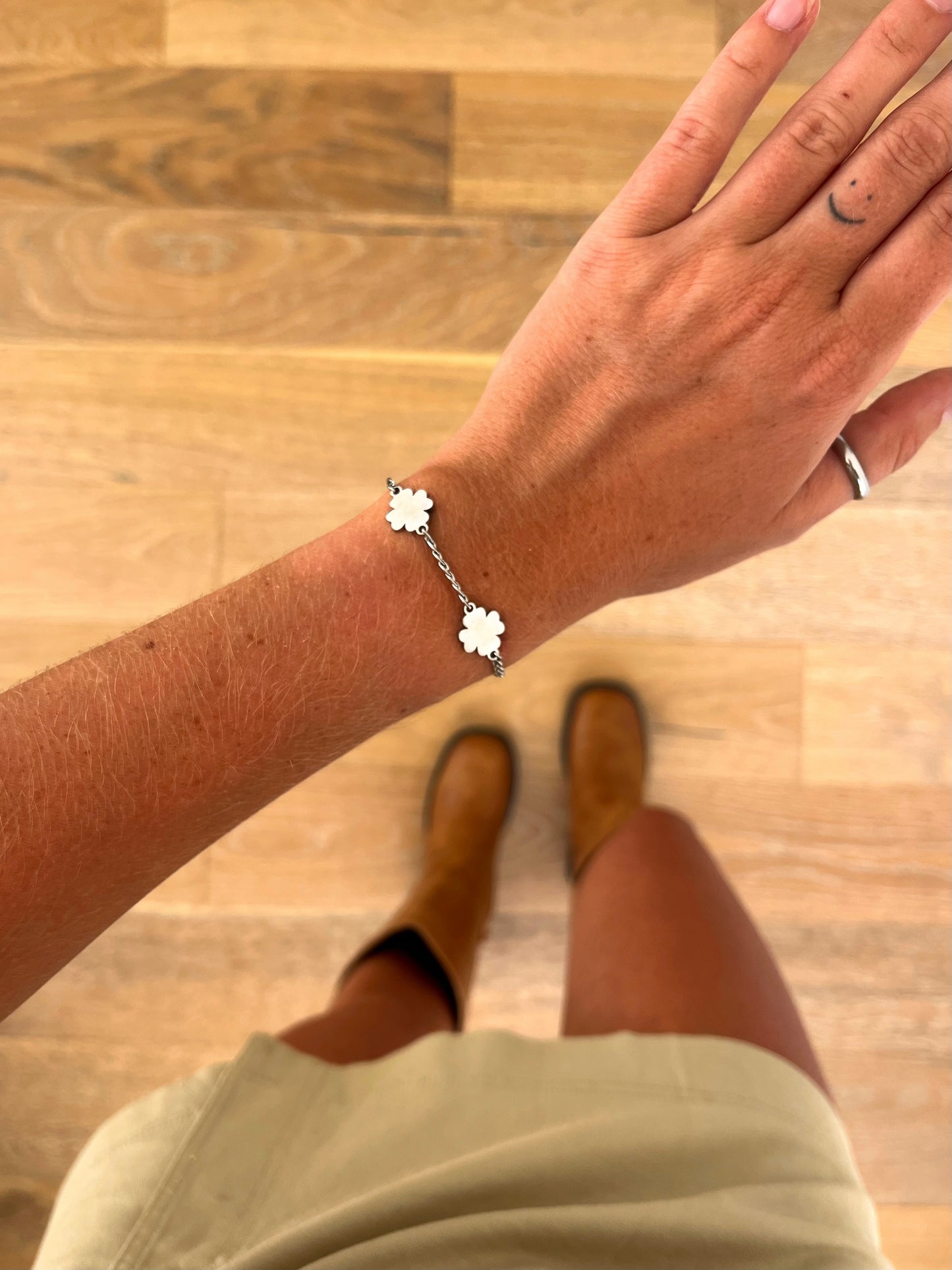 A wrist wearing the Fora I four-leaf clover bracelet in polished stainless steel, photographed from above with a beige skirt and brown boots in the background.