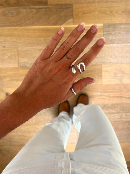 A hand wearing the Kae paperclip chain ring in polished stainless steel, photographed from above with white denim and brown boots in the background.