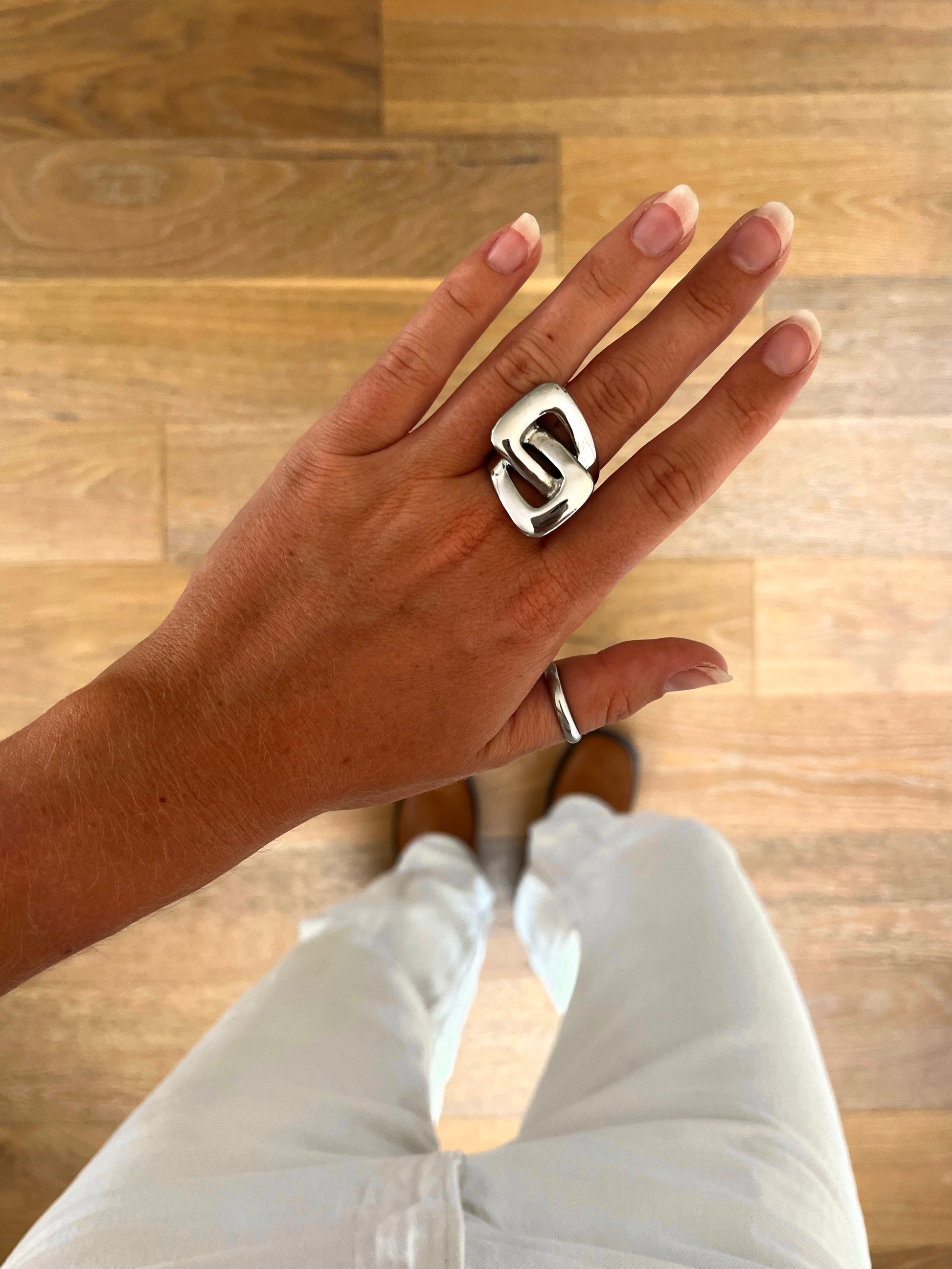 A hand wearing the Kaia sculptural linked ring in polished stainless steel, photographed from above with white denim and brown boots in the background.