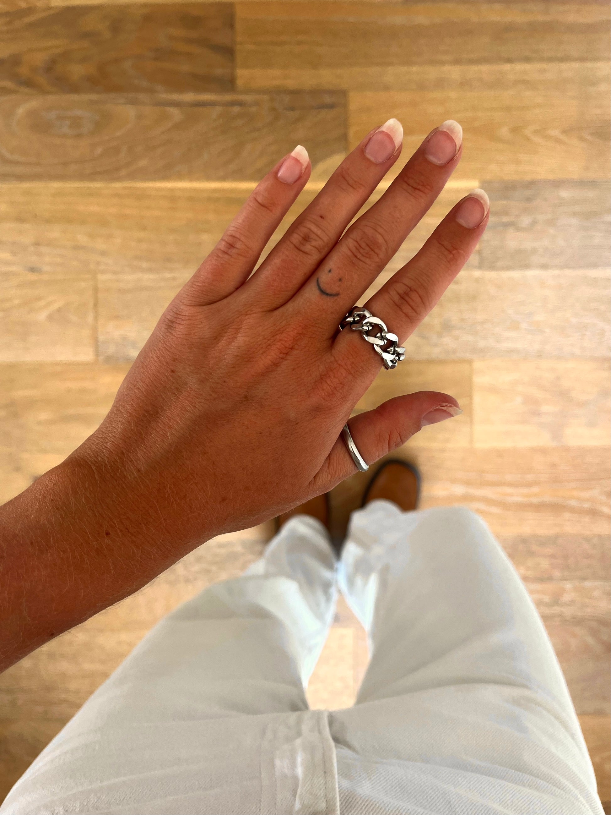A hand wearing the Kairn chain ring in polished stainless steel, photographed from above with white denim and brown boots in the background.