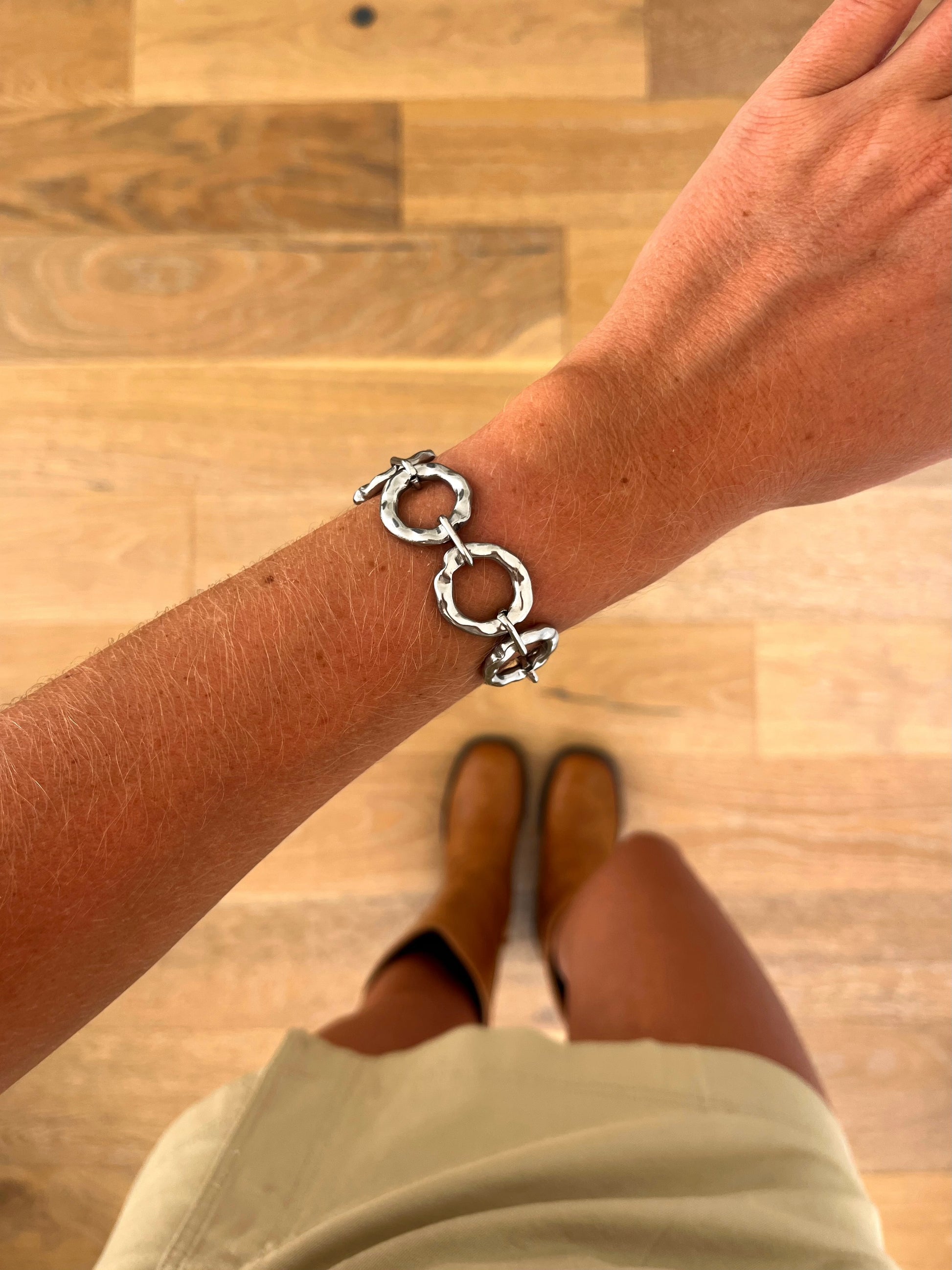 A wrist wearing the Koro sculptural link bracelet in polished stainless steel, photographed from above with a beige skirt and brown boots in the background.