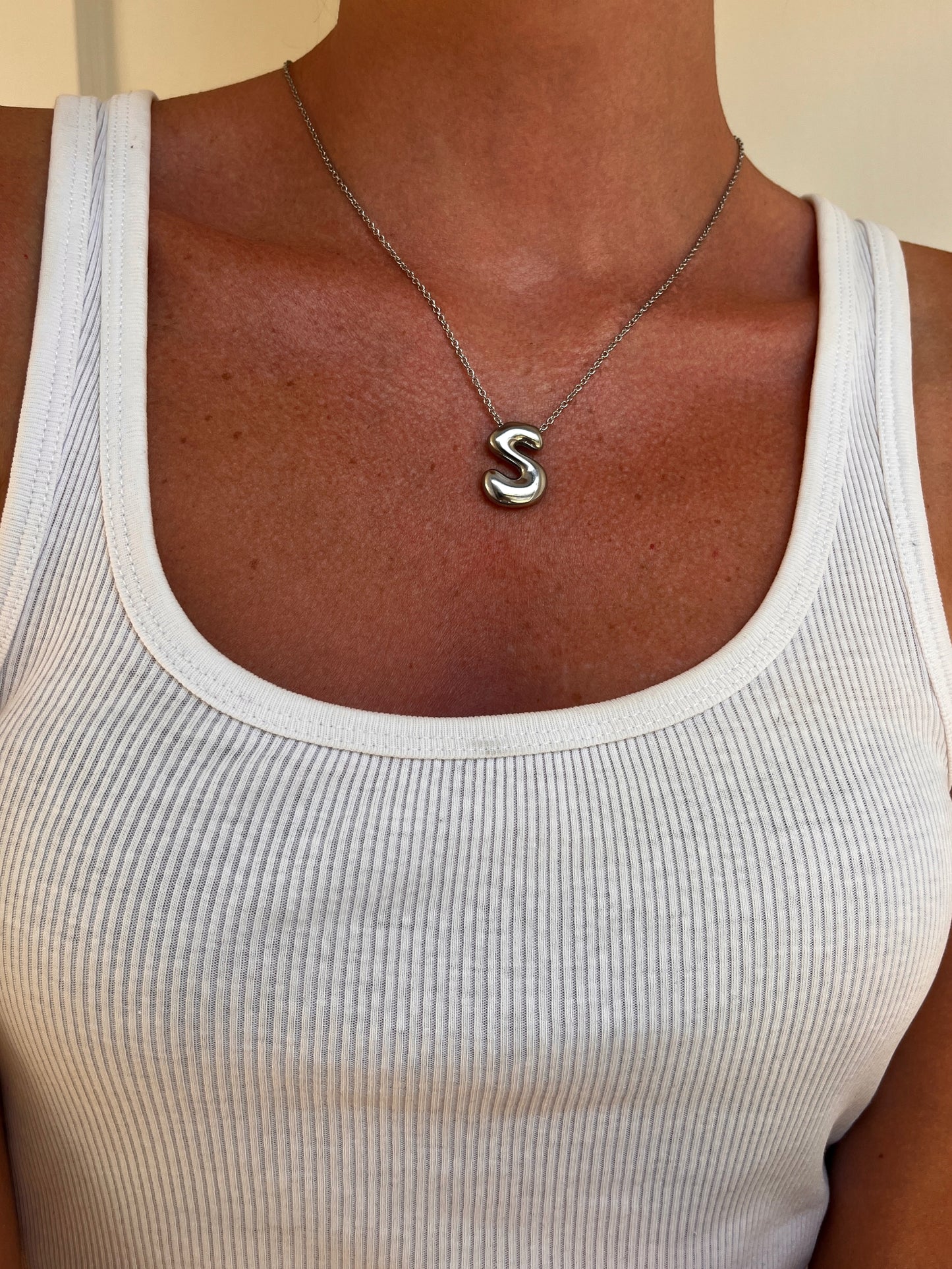 A close-up of a person wearing a silver initial necklace with the letter 'S' pendant, styled with a white ribbed tank top.