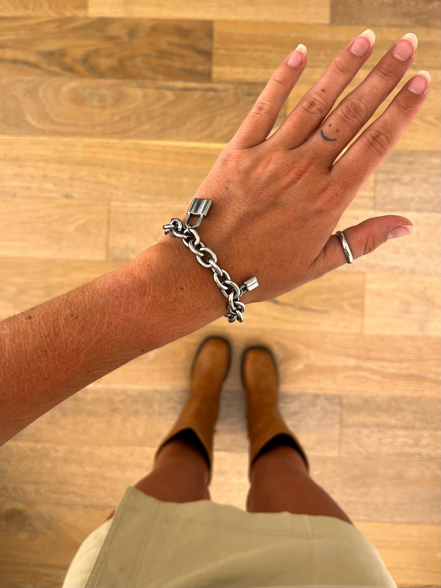 A wrist wearing the Locke sculptural chain bracelet in polished stainless steel, photographed from above with a beige skirt and brown boots in the background.