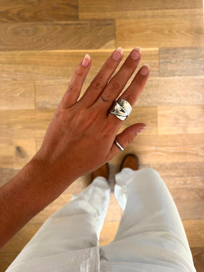 A hand wearing the Mara sculptural wrapped statement ring in polished stainless steel, photographed from above with white denim and brown boots in the background.