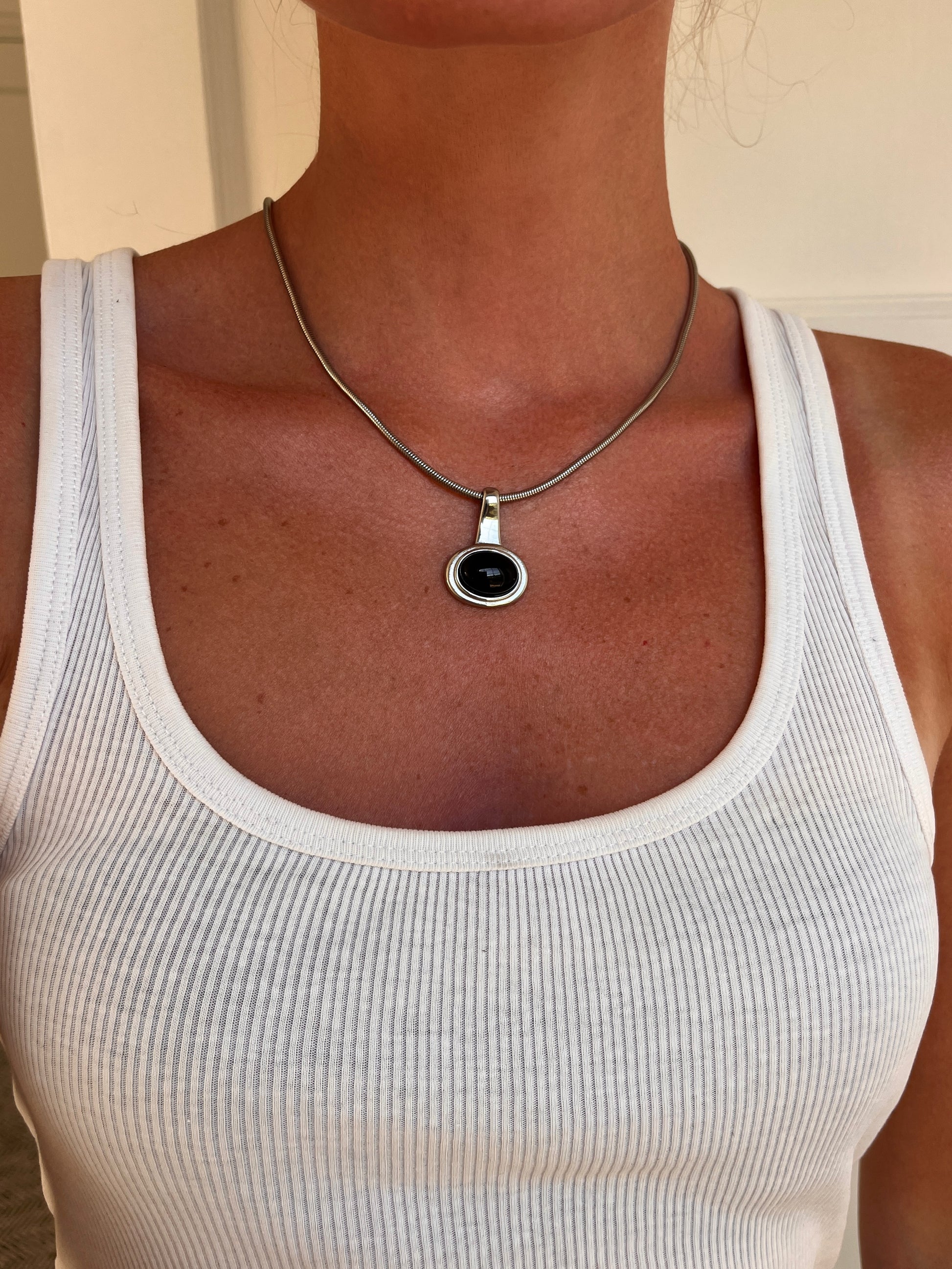 A close-up of a person wearing the medea necklace a silver-toned chain necklace with a round black pendant, paired with a white ribbed tank top.