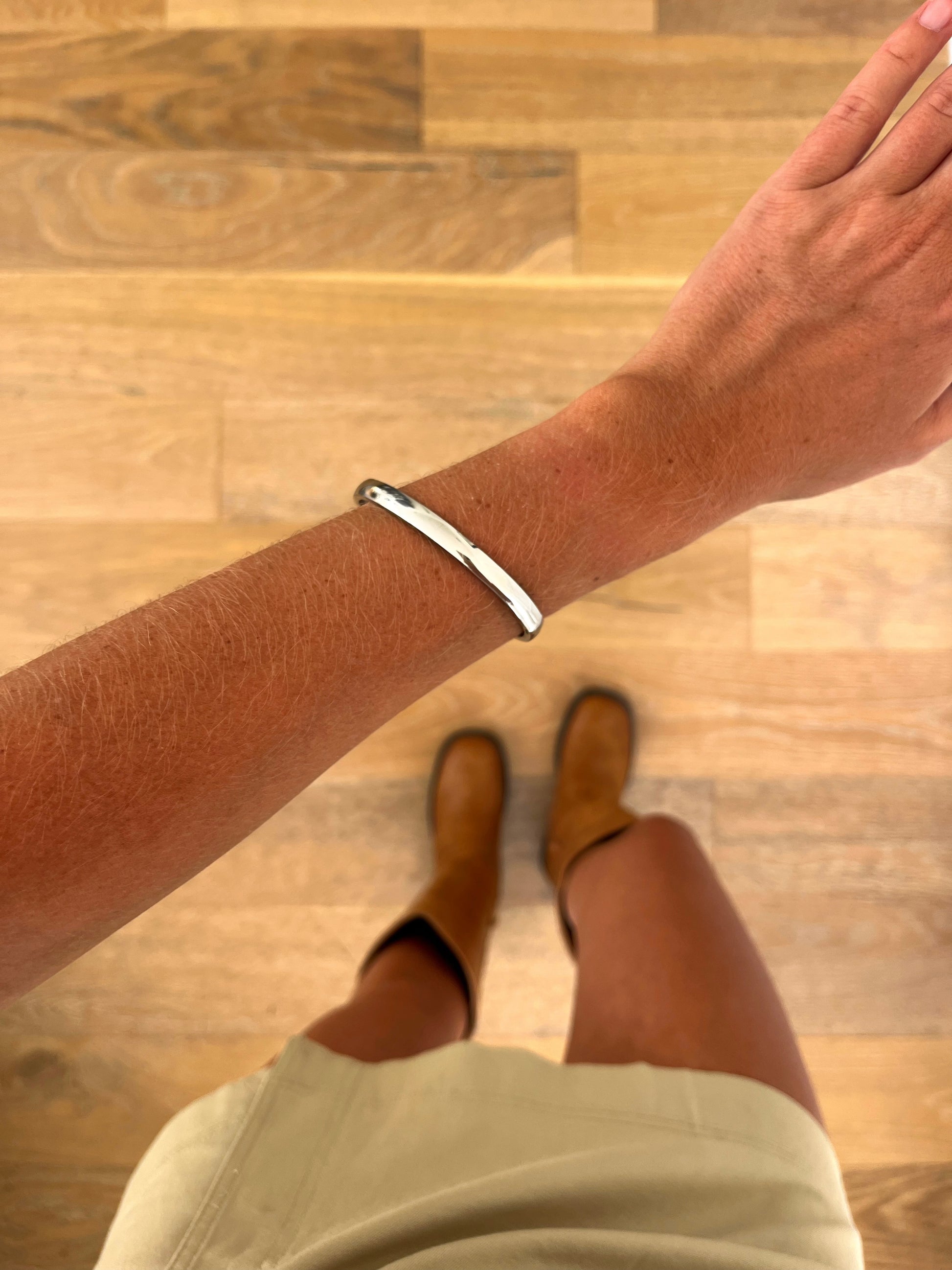 A wrist wearing the Meridian polished stainless steel bangle, photographed from above with a beige skirt and brown boots in the background.