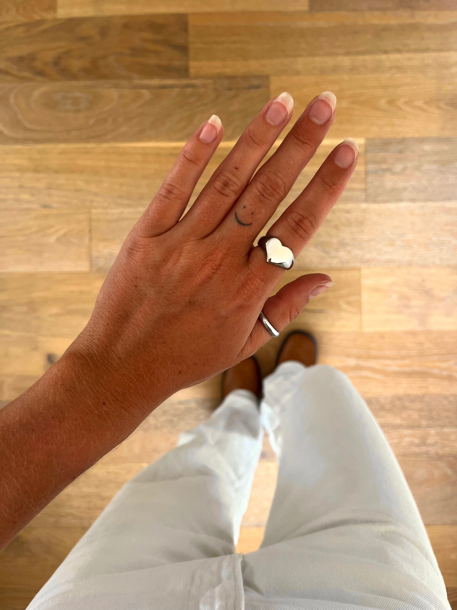A hand wearing the Mora sculptural heart ring in polished stainless steel, photographed from above with white denim and brown boots in the background.