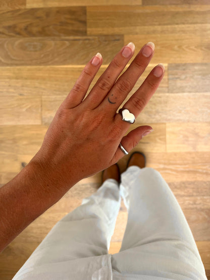 A hand wearing the Mora sculptural heart ring in polished stainless steel, photographed from above with white denim and brown boots in the background.