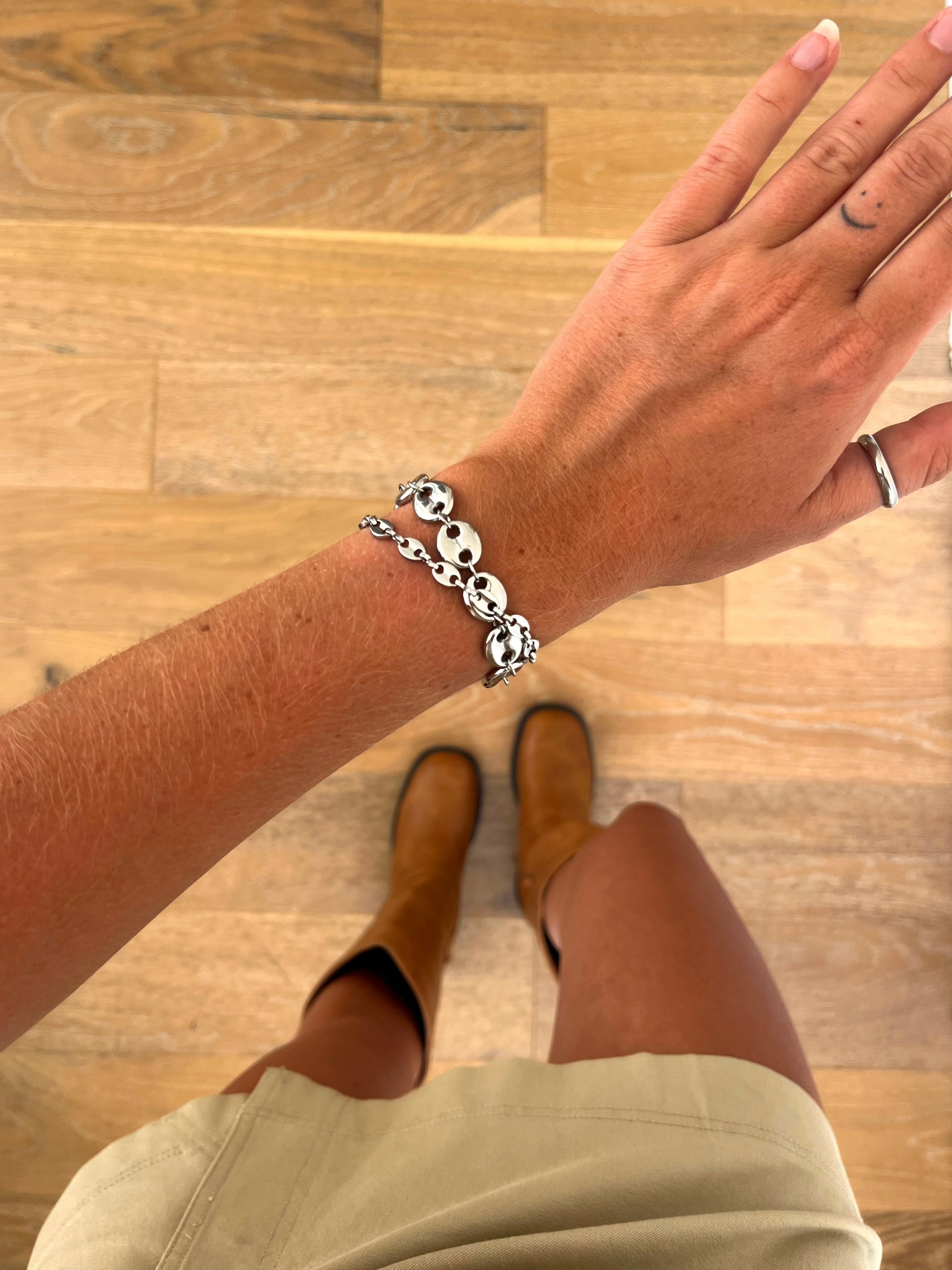 A wrist wearing the Noma I and II sculptural chain bracelets in polished stainless steel, photographed from above with a beige skirt and brown boots in the background.