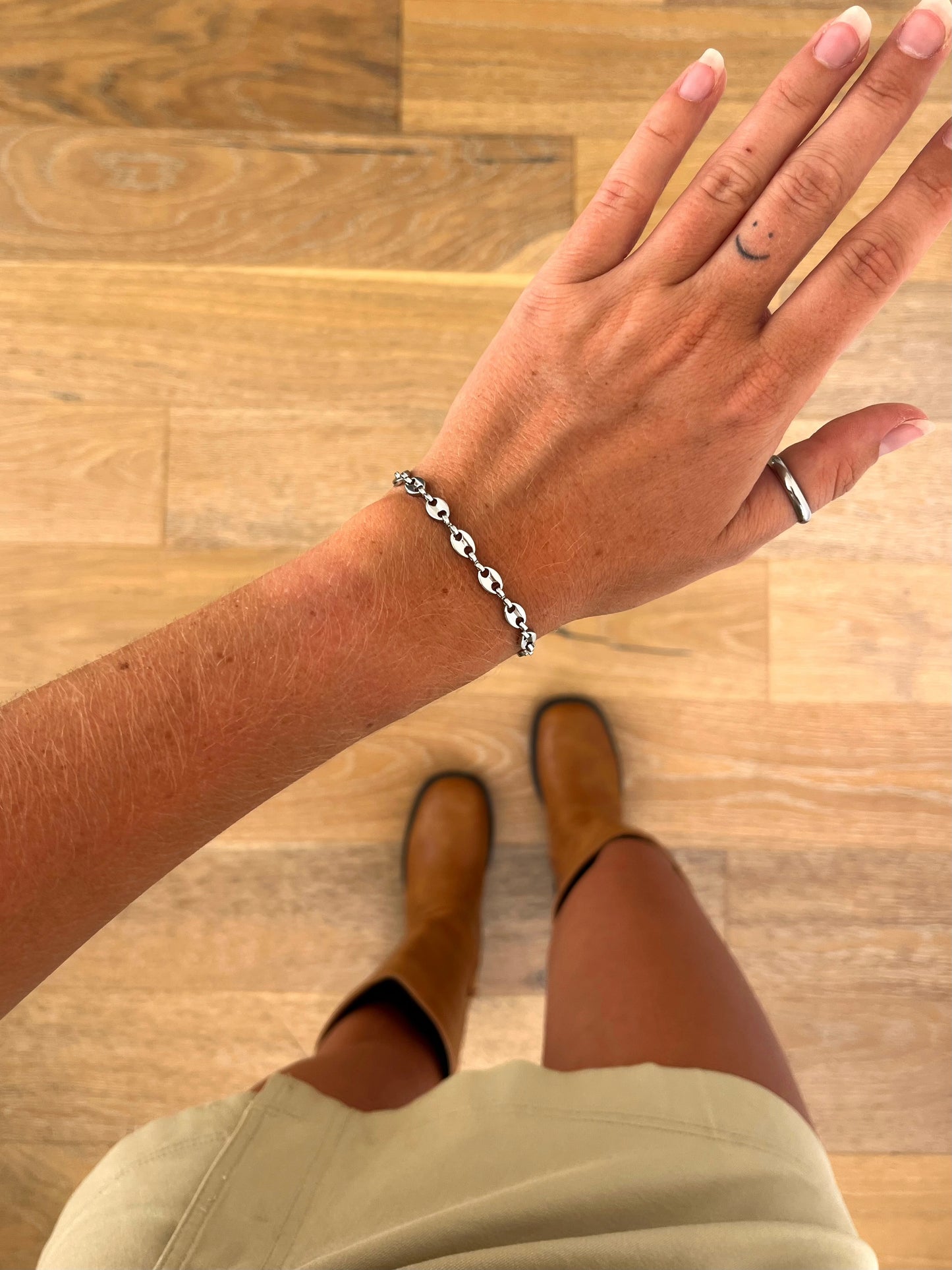 A wrist wearing the Noma I sculptural chain bracelet in polished stainless steel, photographed from above with a beige skirt and brown boots in the background.