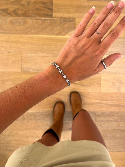 A wrist wearing the Noma I sculptural chain bracelet in polished stainless steel, photographed from above with a beige skirt and brown boots in the background.