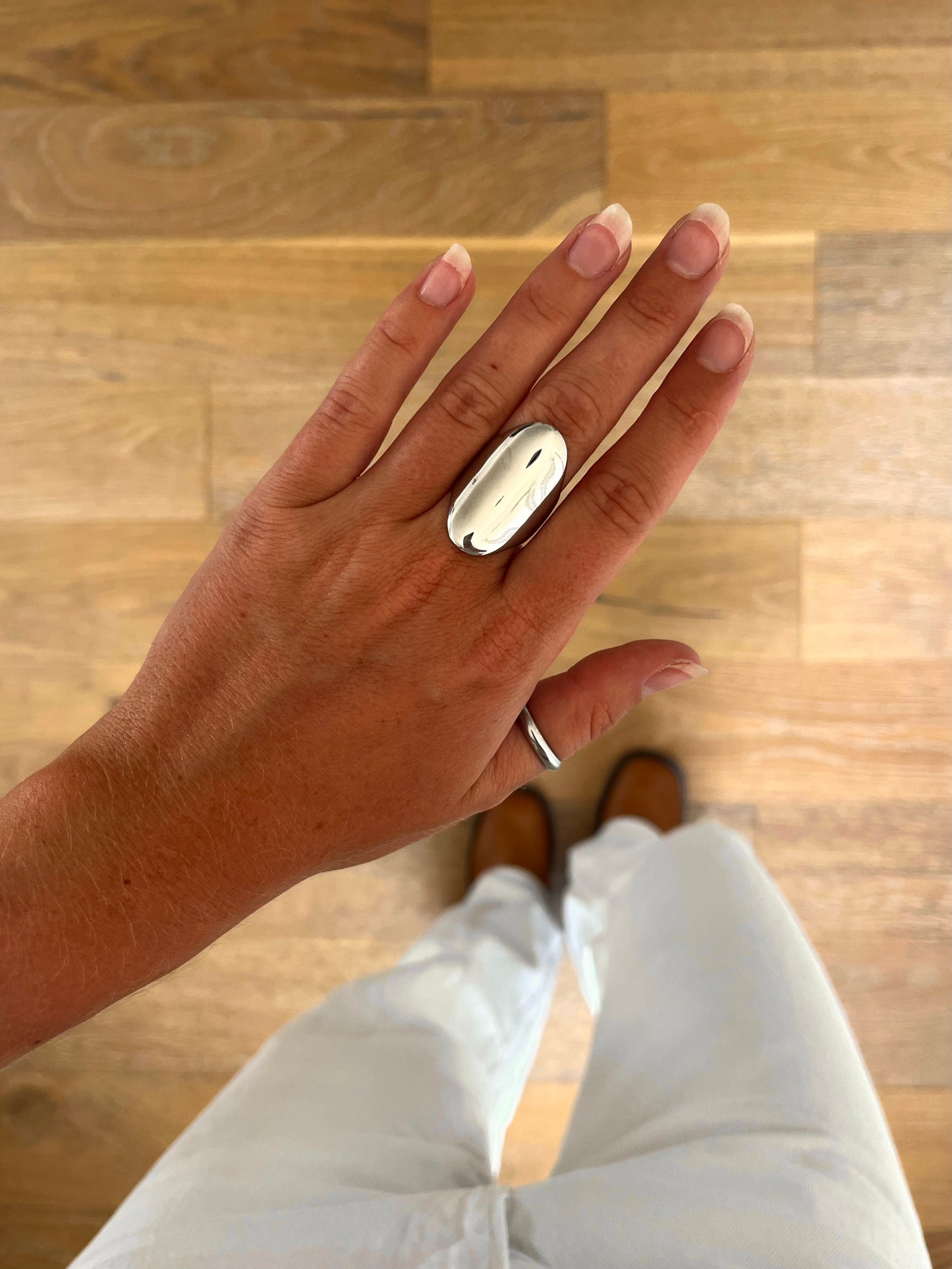 A hand wearing the Ova sculptural oval statement ring in polished stainless steel, photographed from above with white denim and brown boots in the background.