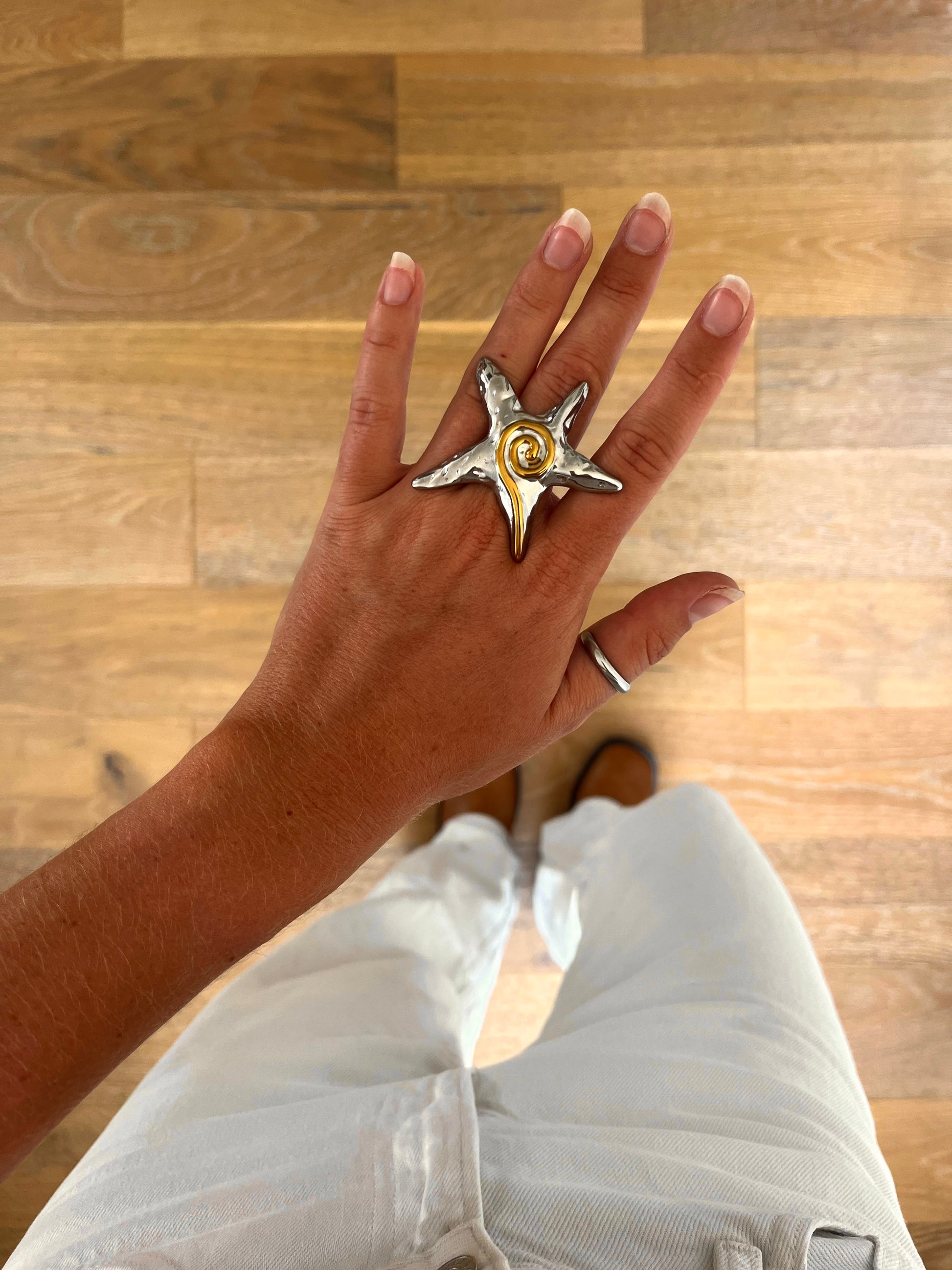 A hand wearing the Solara sculptural starfish ring in polished stainless steel, photographed from above with white denim and brown boots in the background.