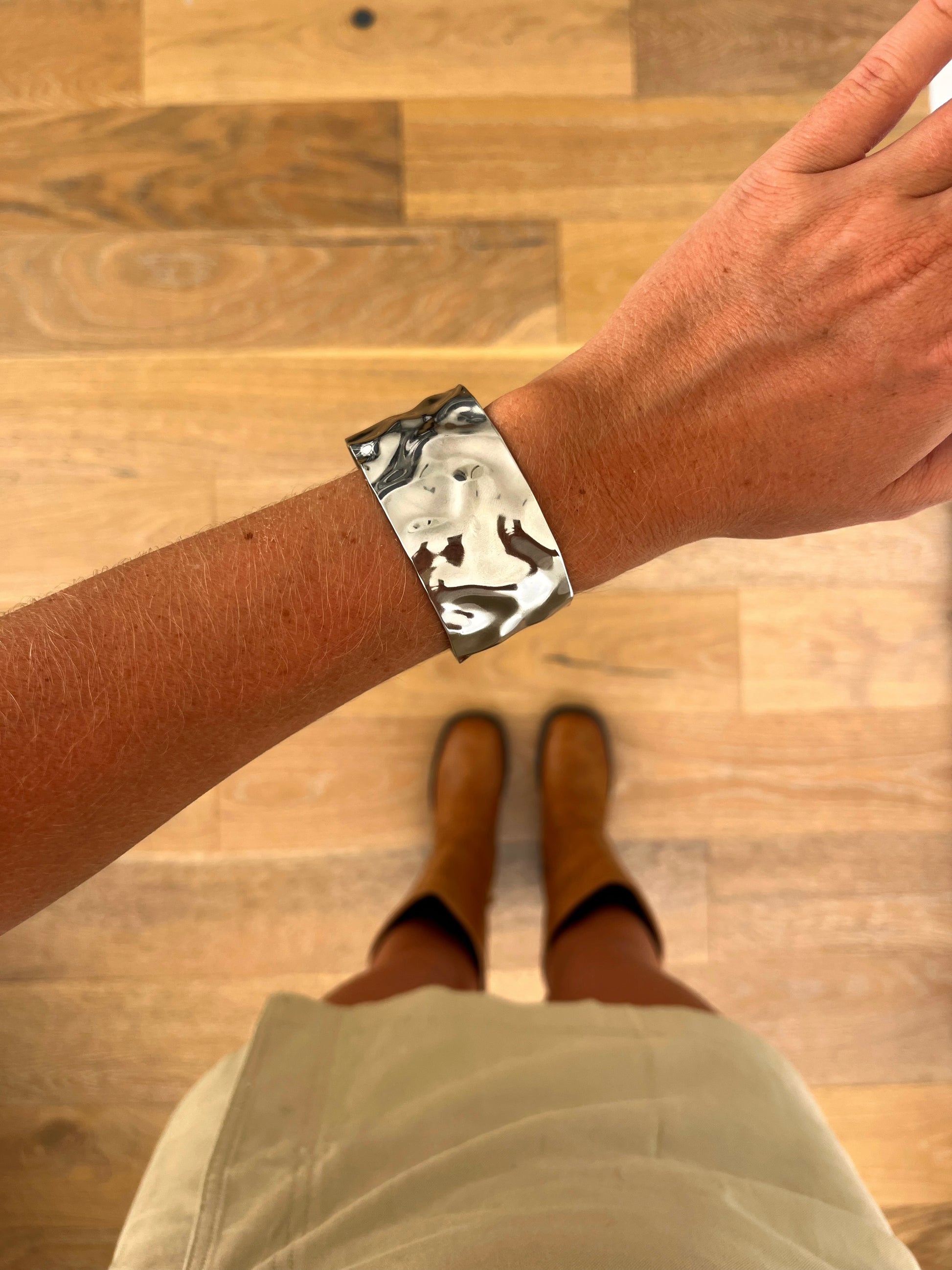 A wrist wearing the Strata textured sculptural stainless steel bangle, photographed from above with a beige skirt and brown boots in the background.