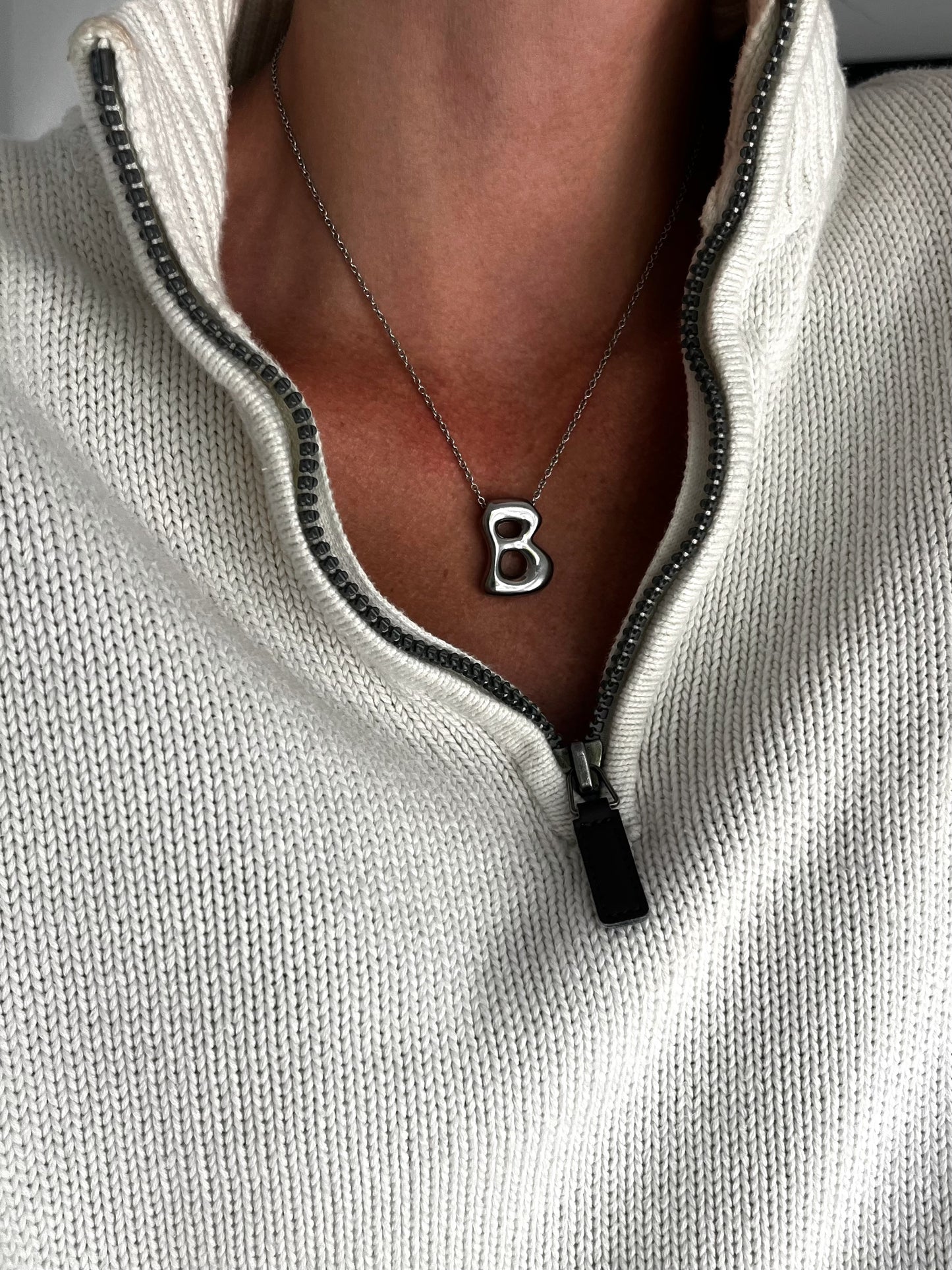 Necklace with a 'B' pendant worn by a person in a light gray sweater.