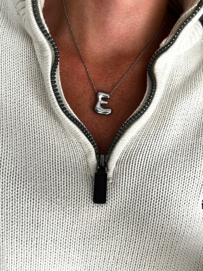 Necklace with a letter 'E' pendant on a person wearing a light gray zip-up hoodie.