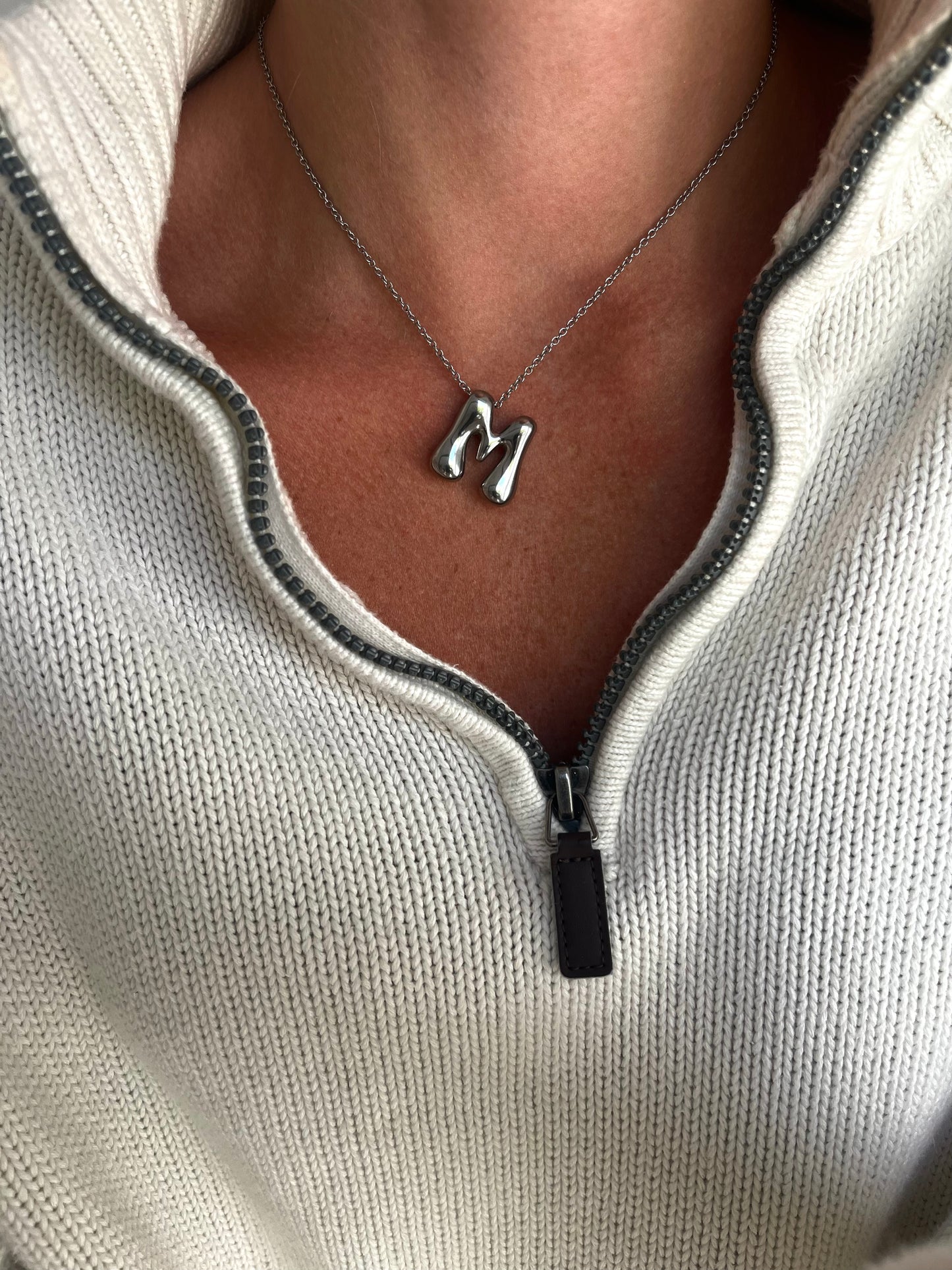 Necklace with a letter 'M' pendant worn by a person in a light-colored sweater.