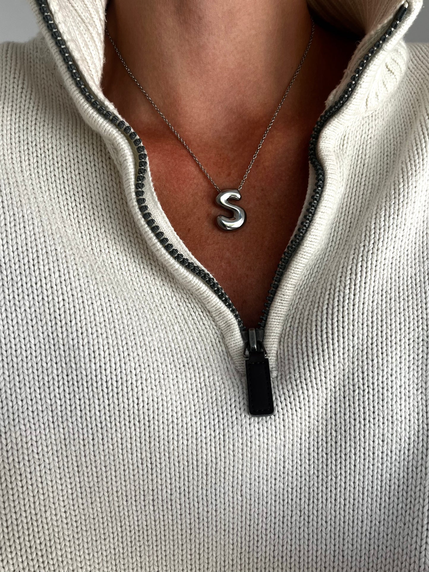Person wearing a light gray zip-up hoodie with a necklace featuring a letter 'S'.