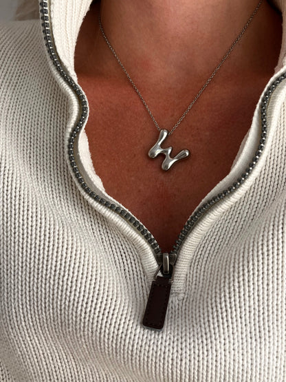 Silver necklace with a unique letter w pendant worn by a person in a light gray sweater.